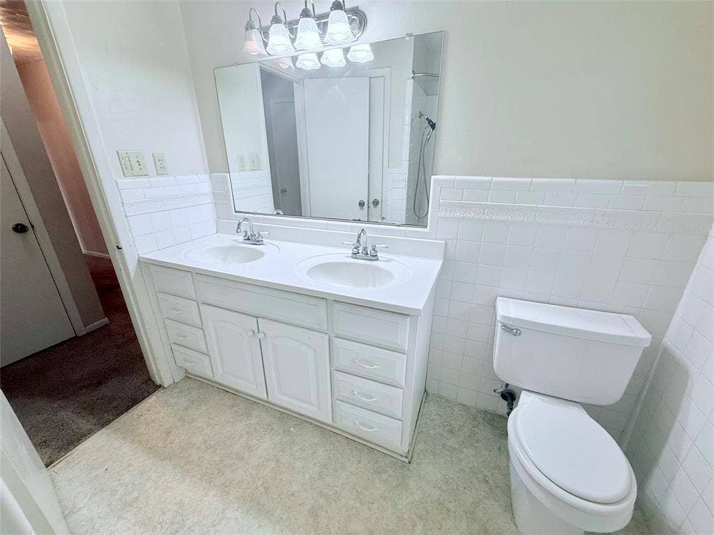 1500 S Houston, Denison, TX 75020 Double-sink vanity featuring a solid surface countertop, white shaker-style cabinetry, a wall-mounted mirror, and tile wainscoting