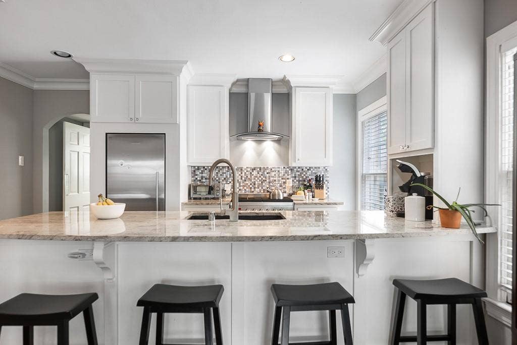 Kitchen featuring white cabinetry, stone countertops, a stainless steel range hood, a mosaic tile backsplash, and a central island with an integrated sink