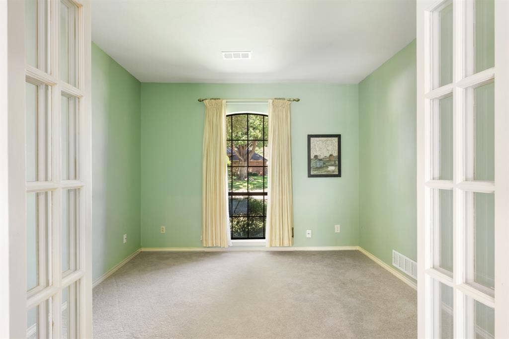 2510 Buttercup Drive, Richardson, TX 75082 Light-filled private office featuring French doors, soft green walls, plush carpeting, and a charming arched window overlooking the front yard. The inviting space offers a peaceful setting for a home office, study, or flex space.