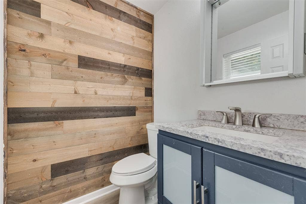 1238 Neptune Drive, Cedar Hill, TX 75104 Bathroom featuring a multi-tone wood-plank accent wall, a blue vanity with frosted glass cabinet inserts, a stone-composite countertop, and a built-in medicine cabinet with integrated lighting