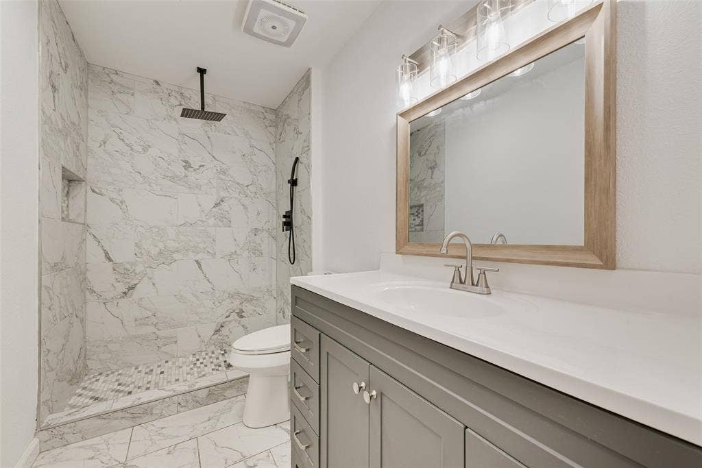 1238 Neptune Drive, Cedar Hill, TX 75104 Bathroom featuring a walk-in shower with stone-pattern tile surround, recessed wall niche, matte black rainfall showerhead, gray shaker-style vanity with white solid-surface countertop, and wood-framed mirror