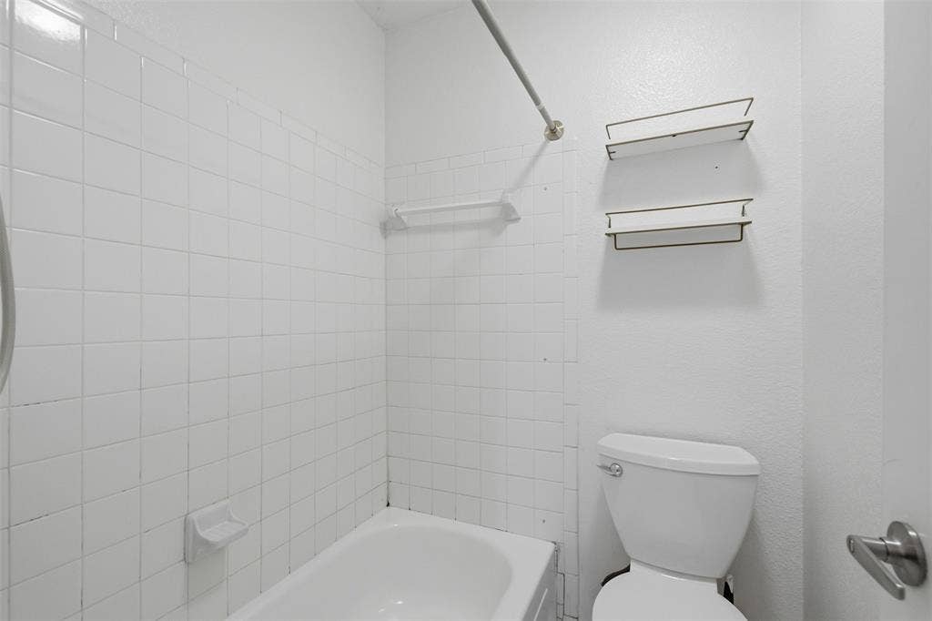 Full-height white square tile tub surround, built-in soap dish, shower curtain rod, wall-mounted storage shelving, and standard porcelain toilet