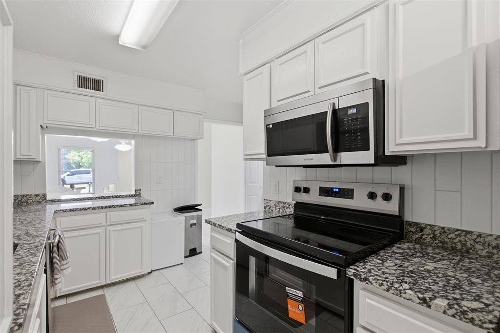 Kitchen featuring white shaker-style cabinetry, speckled stone-finish countertops, stainless steel appliances, vertical tile backsplash, and tile flooring