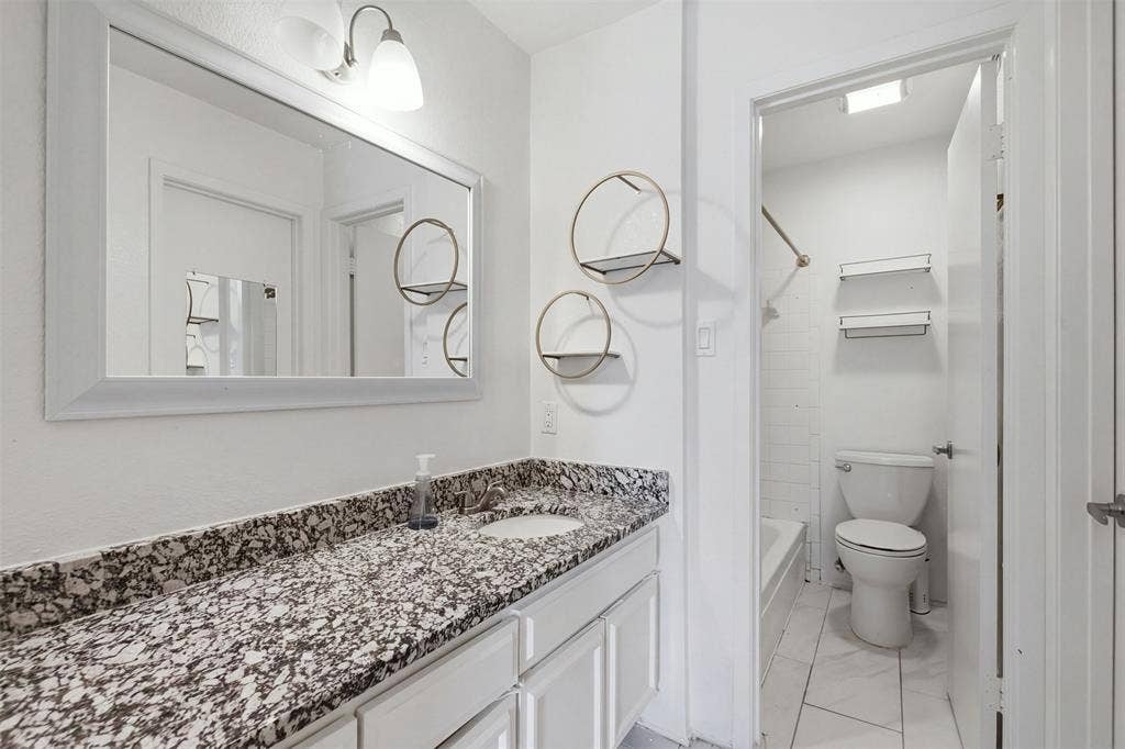 Bathroom featuring a vanity with a stone-patterned countertop, an integrated sink, a framed wall mirror, dual-light wall-mounted fixture, and a tiled shower-tub combination