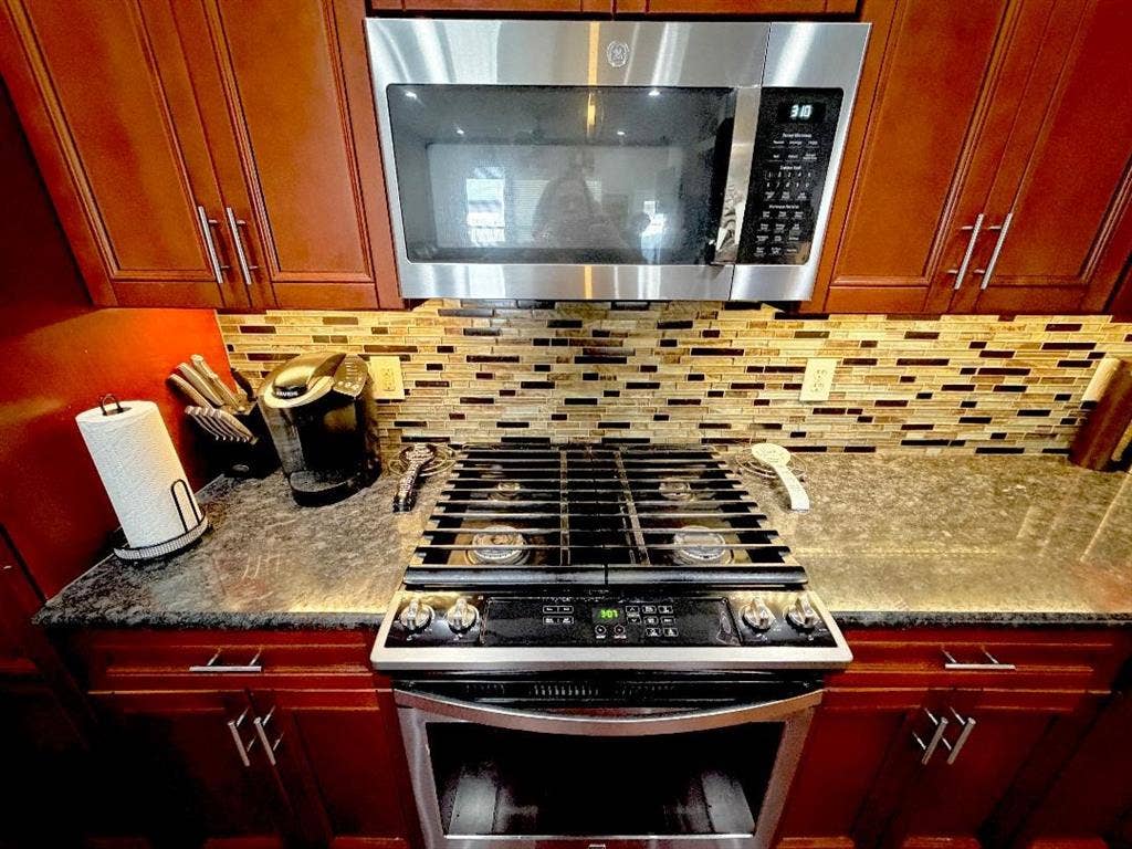 Kitchen featuring wood cabinetry, natural stone countertops, a multi-tone glass tile backsplash, and stainless steel gas range with a matching over-the-range microwave