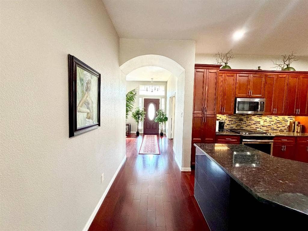 Kitchen featuring wood cabinetry, stone countertops, and a tiled backsplash, leading to an entryway with an arched transition and wood-finish flooring