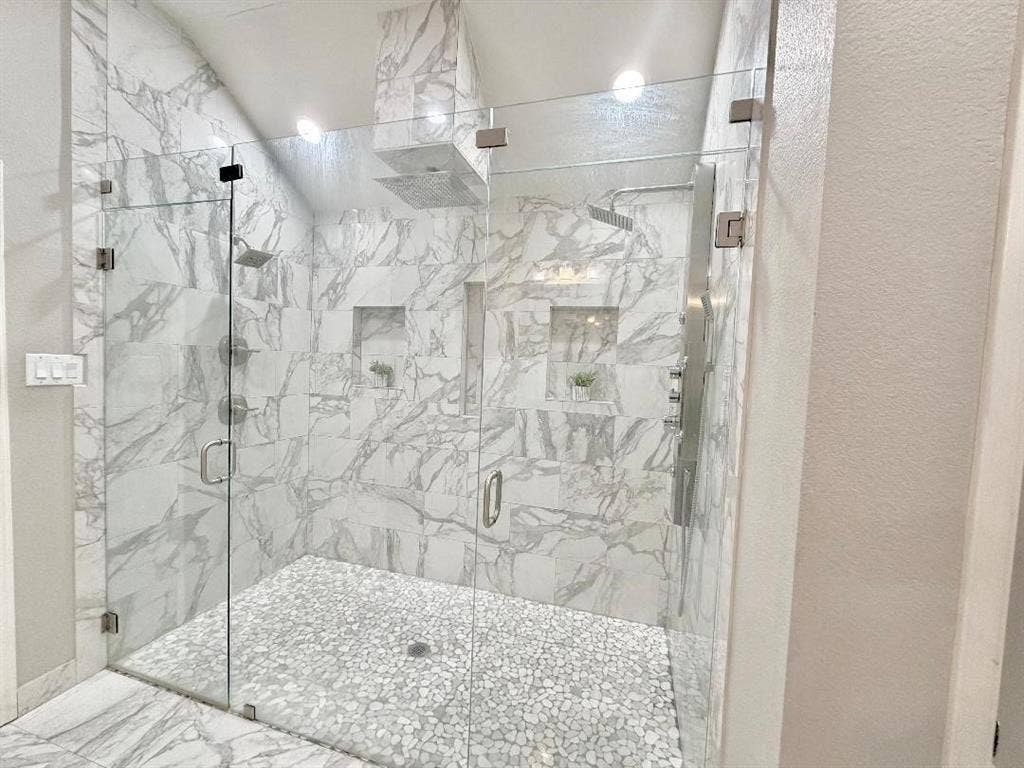Walk-in shower featuring floor-to-ceiling stone-pattern tile, frameless glass enclosure, pebble-stone flooring, dual recessed wall niches, and a multi-head shower system