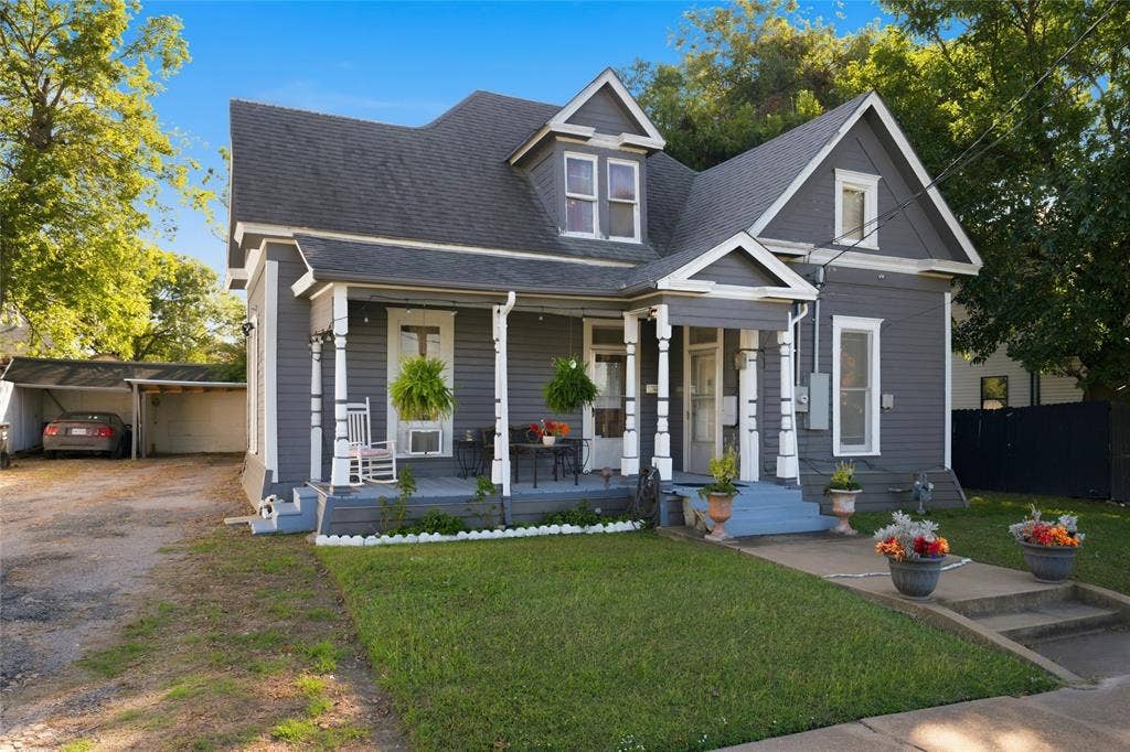 2309 Jones Street, Greenville, TX 75401 Victorian-style exterior featuring gray horizontal siding, a covered front porch with decorative turned posts, multiple roof gables, and a detached carport