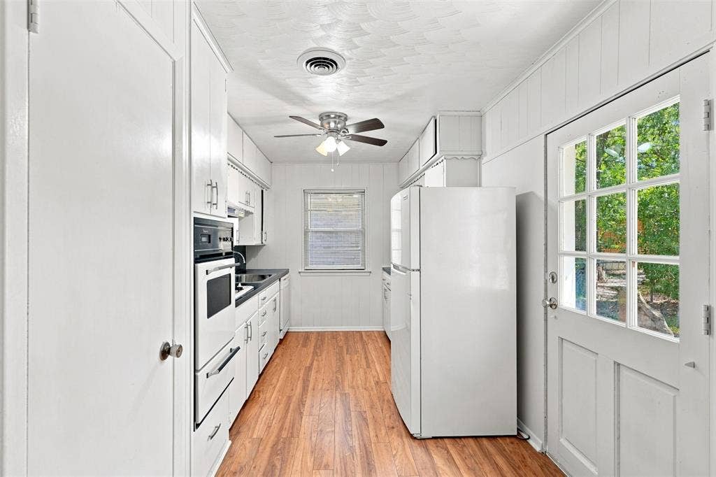 Galley kitchen featuring wood-finish flooring, white cabinetry with contemporary hardware, integrated wall oven, and a paneled glass-insert door