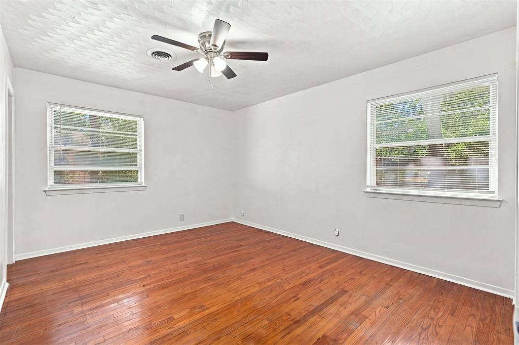 Room featuring rich wood-finish flooring, two windows with blinds, a ceiling fan with integrated lighting, a ceiling vent, and light-toned wall finishes