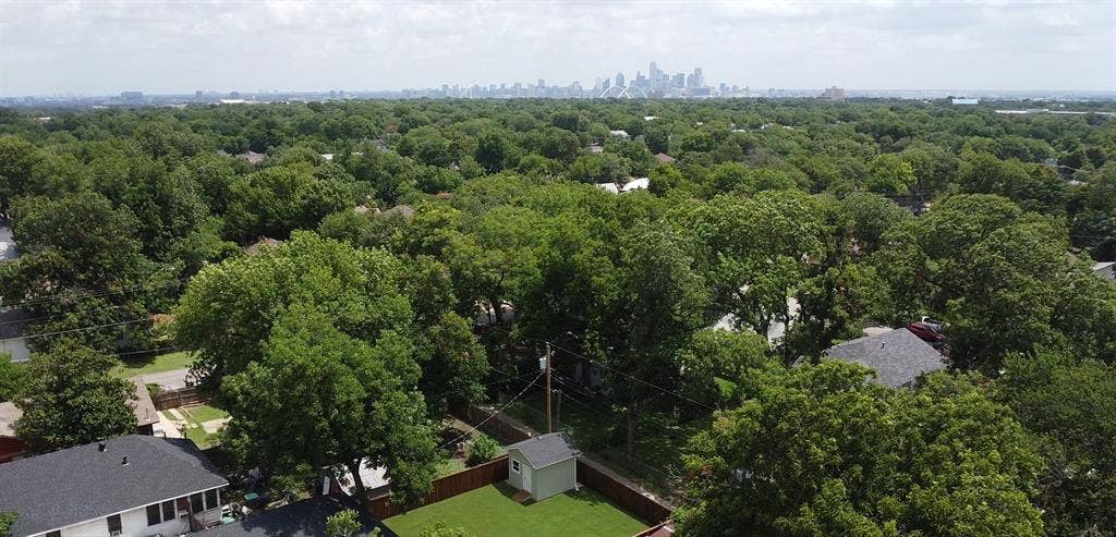 922 Hollywood Avenue, Dallas, TX 75208 Aerial perspective featuring a backyard with a detached storage structure, enclosed by fencing, set against a backdrop of mature tree canopy and a city skyline