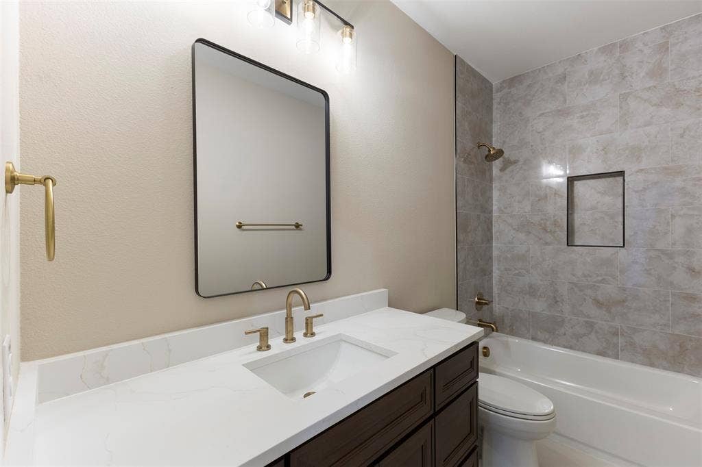 2390 Usman Drive, Irving, TX 75062 Bathroom featuring a dark wood-finish vanity with a white countertop, rectangular undermount sink, brass-toned fixtures, a framed wall mirror, and a tub with tiled surround and built-in recessed niche