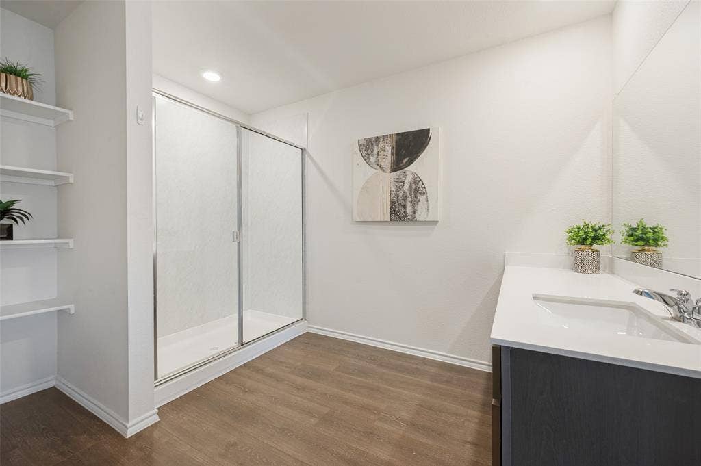 Shower enclosure with metal framing, vanity featuring a solid surface countertop, built-in shelving, full-width vanity mirror, and wood-finish flooring