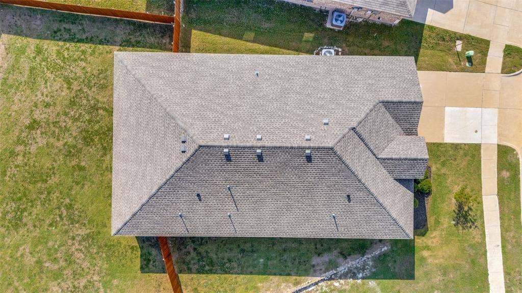 Aerial perspective featuring a multi-pitch asphalt shingle roof, concrete driveway, and expansive lawn area enclosed by wood fencing