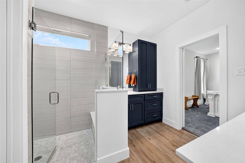 Walk-in shower with floor-to-ceiling tile, glass enclosure, and transom window, paired with a navy-finish vanity featuring quartz countertops and wood-finish flooring