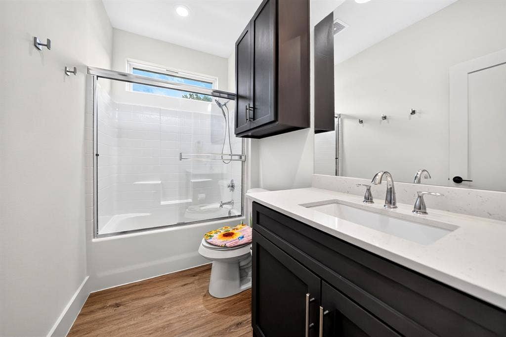Bathroom featuring a dark-finish vanity with a light-toned countertop, integrated sink, shower-tub combination with a glass enclosure, and wood-finish flooring