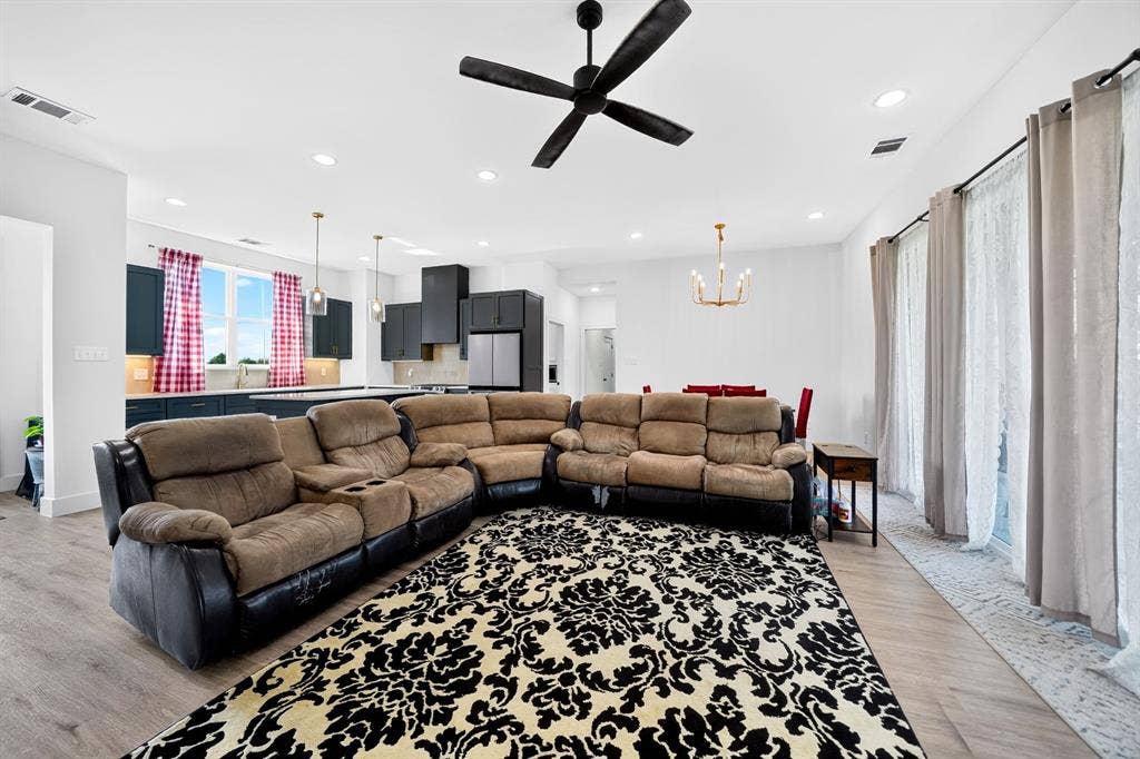 Open-concept living space featuring wood-finish flooring, recessed lighting, matte black ceiling fan, and a kitchen with dark cabinetry and dual pendant lights