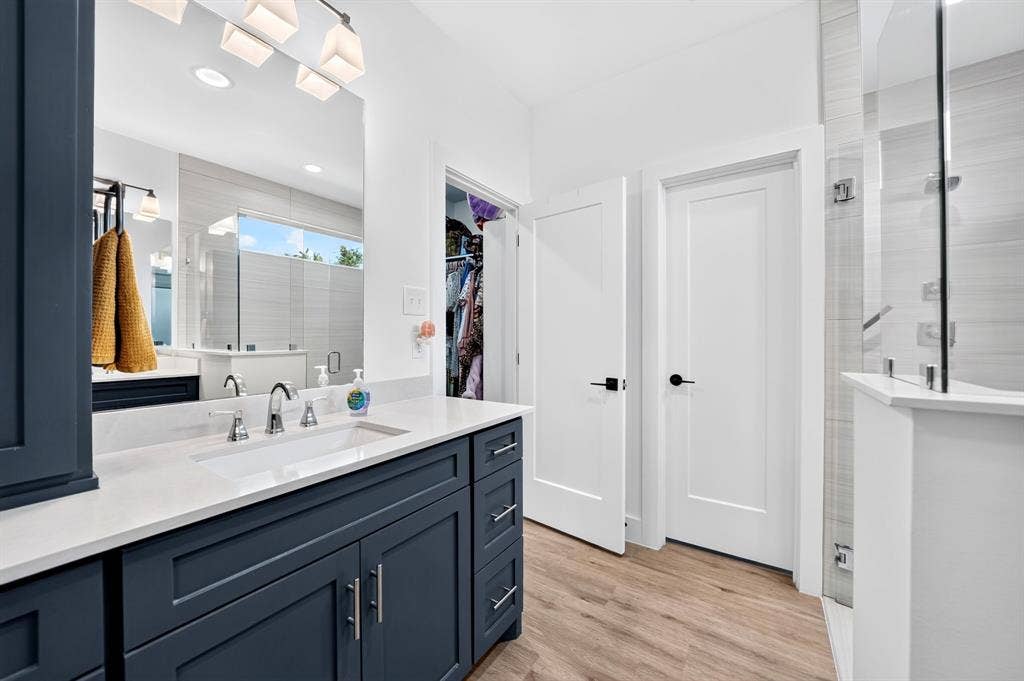 Dark-finish cabinetry paired with a light stone-surface vanity top, integrated rectangular sink, wall-spanning mirror with contemporary multi-light fixture, and walk-in glass-enclosed shower with horizontal-pattern tile surround
