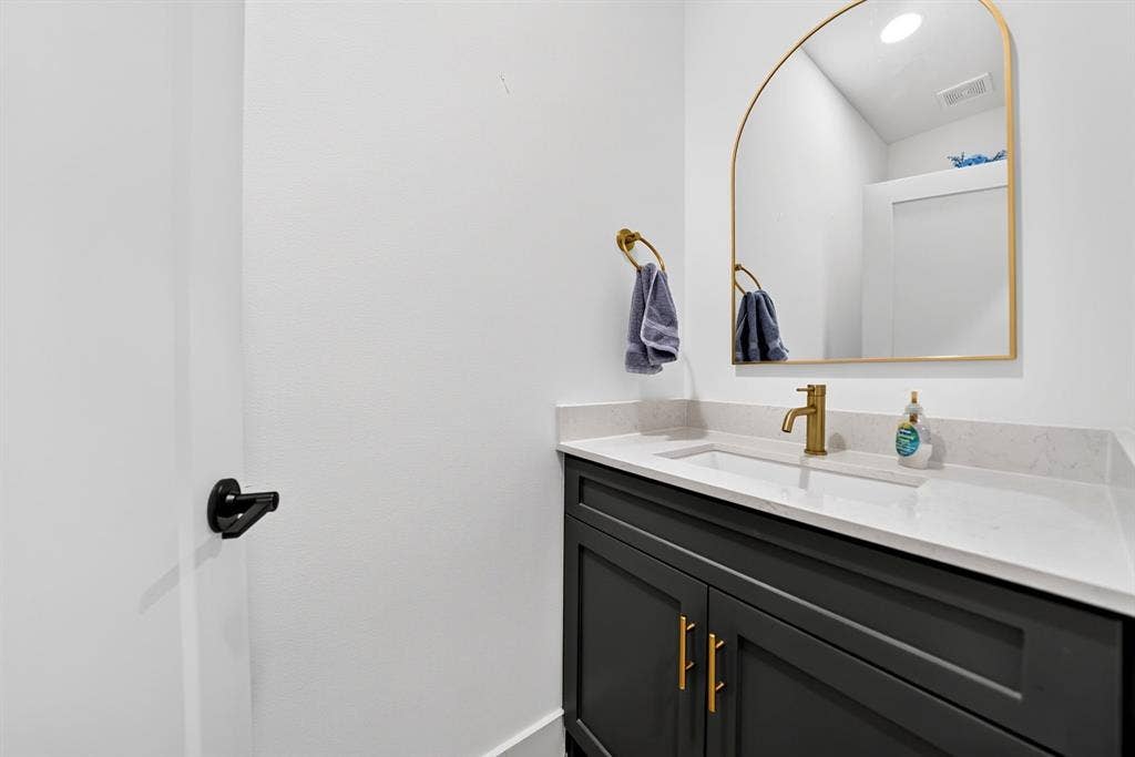 Dark-toned cabinetry featuring gold-finish hardware, quartz countertop, integrated rectangular sink, brass-tone faucet, and arched mirror