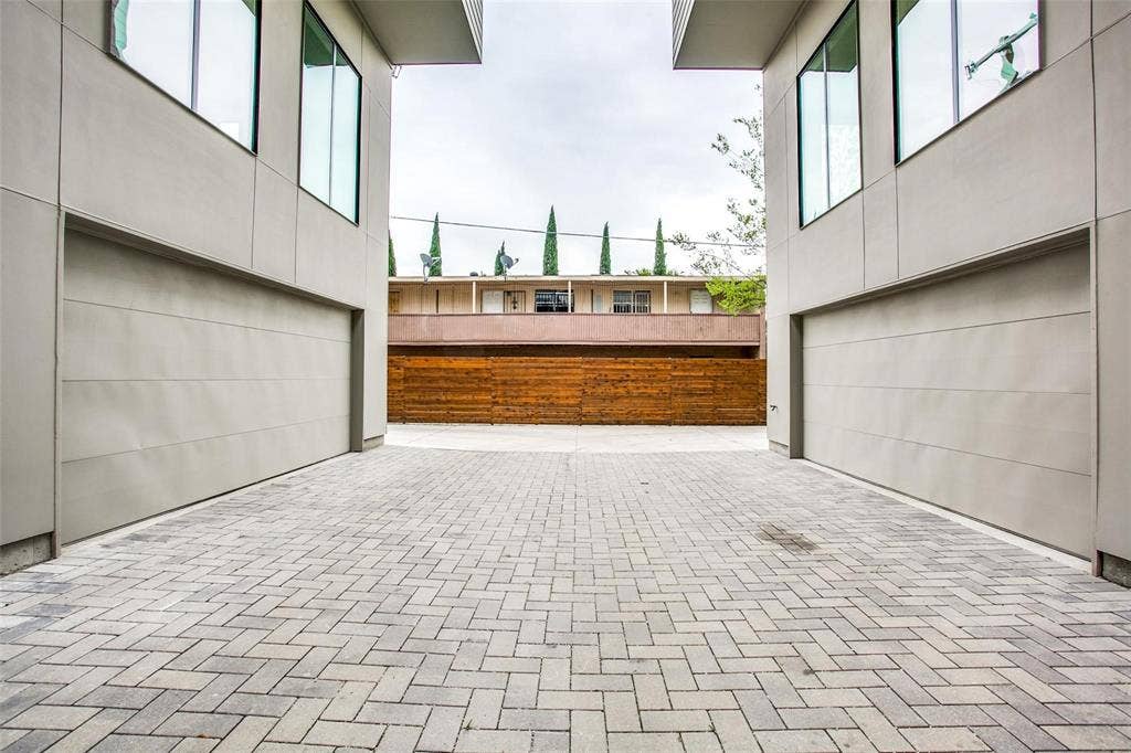 1502 Bennett Avenue, #106, Dallas, TX 75206 Modern exterior facade featuring smooth-finish cladding, dual flush-mount garage doors, and herringbone-pattern paver motor court enclosed by a horizontal wood privacy fence