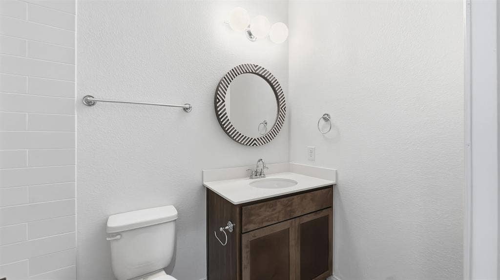 3129 Grayslick Avenue, Celina, TX 75009 Vanity featuring wood-finish cabinetry, a white countertop with an integrated basin, a three-light globe vanity fixture, and a circular decorative wall mirror