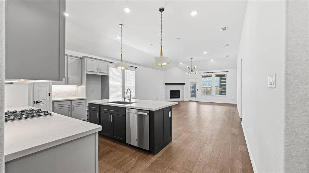 3129 Grayslick Avenue, Celina, TX 75009 Kitchen featuring a large center island with a quartz countertop, contrasting dual-tone cabinetry, stainless steel appliances, and wood-finish flooring throughout the open-concept space, highlighted by a corner fireplace