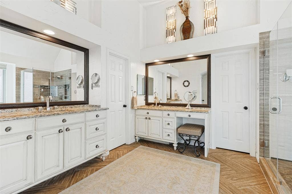 2404 Creek Ridge Drive, McKinney, TX 75072 Dual white cabinetry vanities featuring stone-finish countertops, framed mirrors, and herringbone-patterned flooring, complemented by a glass-enclosed shower