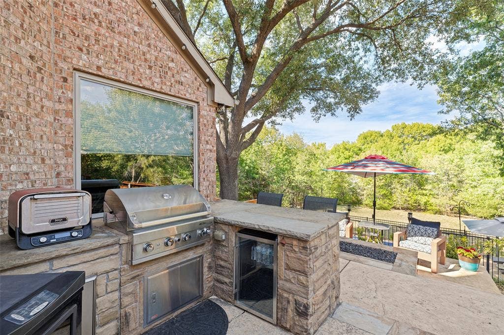 2404 Creek Ridge Drive, McKinney, TX 75072 Outdoor kitchen featuring stone masonry, stainless steel built-in grill, under-counter refrigeration, and stone countertops, set against a backdrop of mature trees