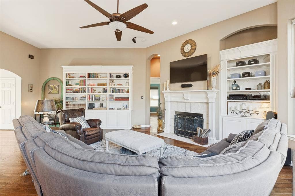 2404 Creek Ridge Drive, McKinney, TX 75072 Living area featuring wood-finish flooring, a fireplace with a white mantel, built-in shelving, arched doorways, and a ceiling fan
