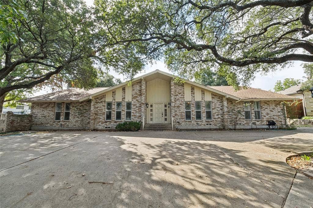 6936 Wildglen Drive, Dallas, TX 75230 Brick exterior featuring a multi-gabled roofline, expansive paved driveway, and mature oak tree canopy