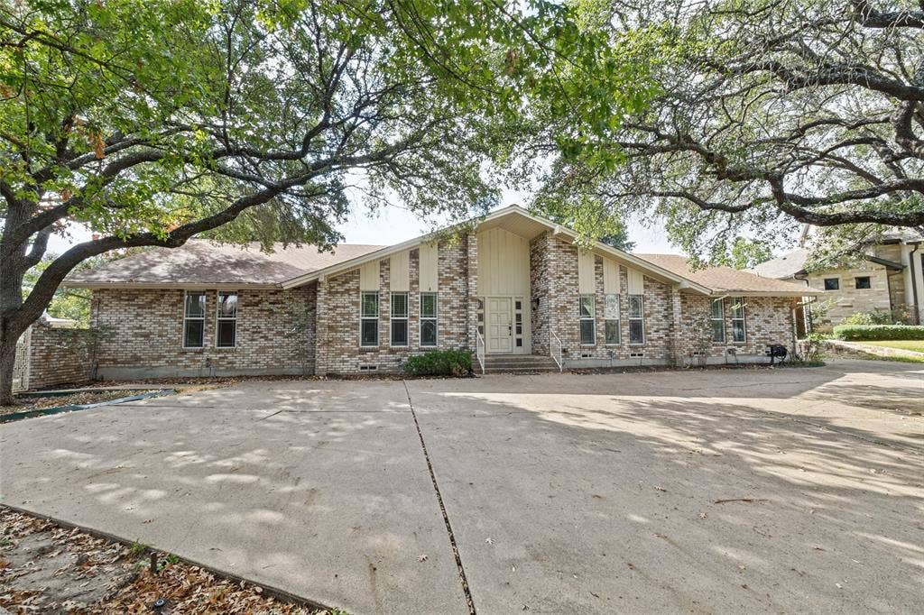 6936 Wildglen Drive, Dallas, TX 75230 Single-story brick exterior featuring a multi-gabled roofline, circle driveway, and mature canopy trees