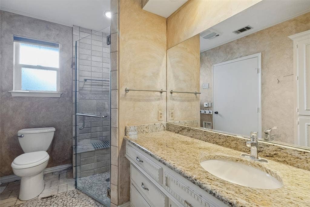 6936 Wildglen Drive, Dallas, TX 75230 Vanity featuring granite countertop, undermount sink, and white cabinetry, complemented by a walk-in shower with glass enclosure and pebble-stone flooring