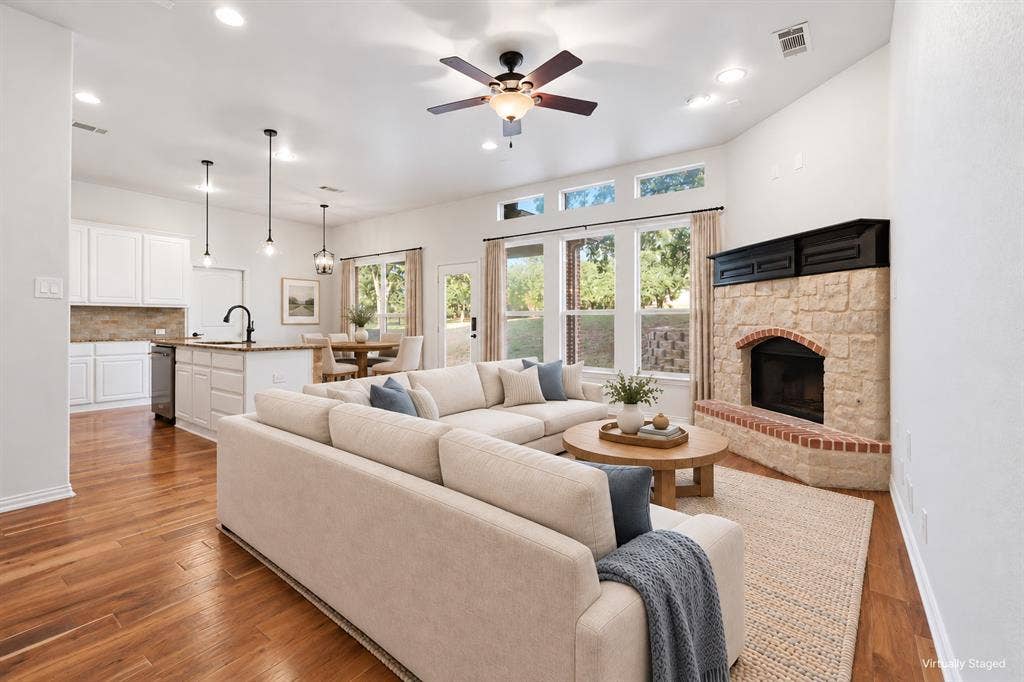 Open floor plan featuring wood-finish flooring, white shaker-style cabinetry, an island with pendant lighting, and a stone fireplace with a brick hearth