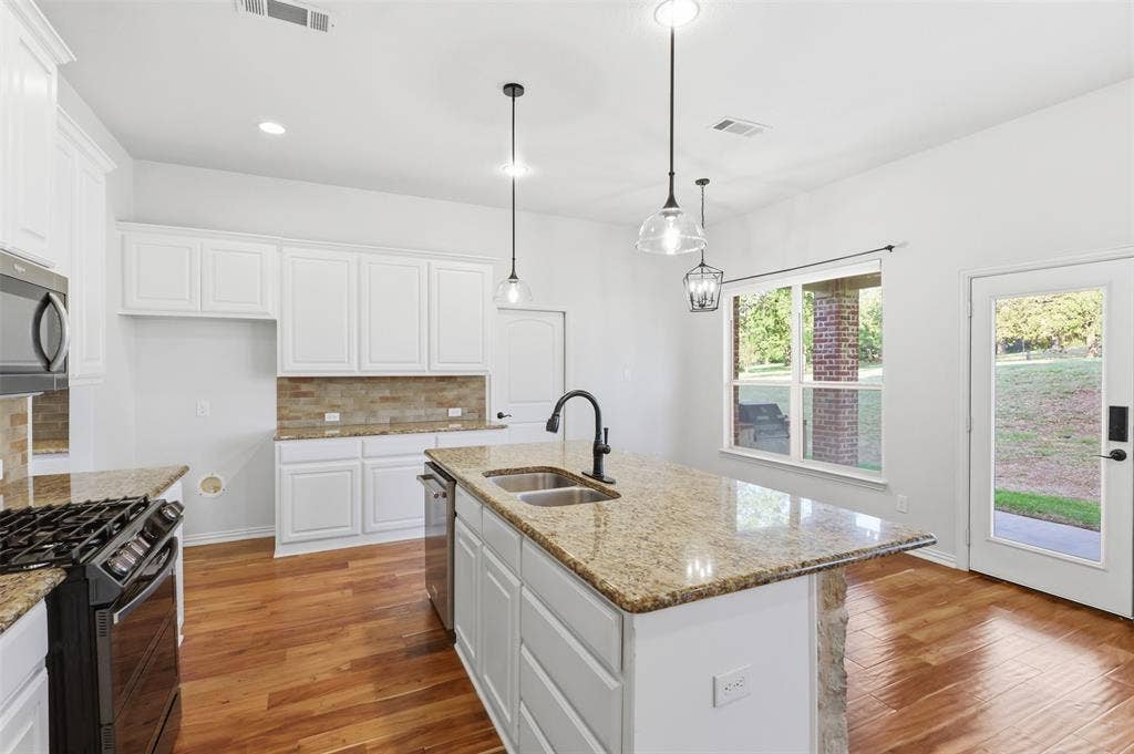 Kitchen featuring white cabinetry, stone-finish countertops, a central island with a dual-basin undermount sink, pendant lighting, and wood-finish flooring
