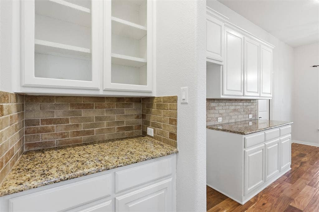Built-in cabinetry featuring upper glass-front doors, stone-patterned countertops, brick-style tile backsplash, and wood-finish flooring