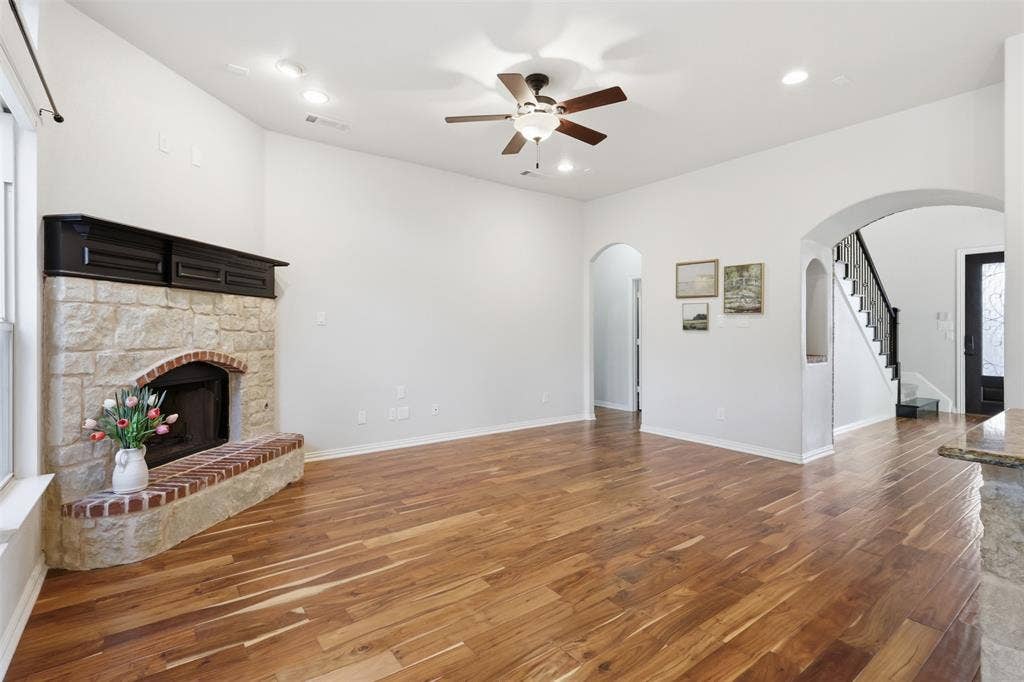 Living area featuring wood-finish flooring, a stone fireplace with a brick hearth, a ceiling fan, and arched architectural entryways