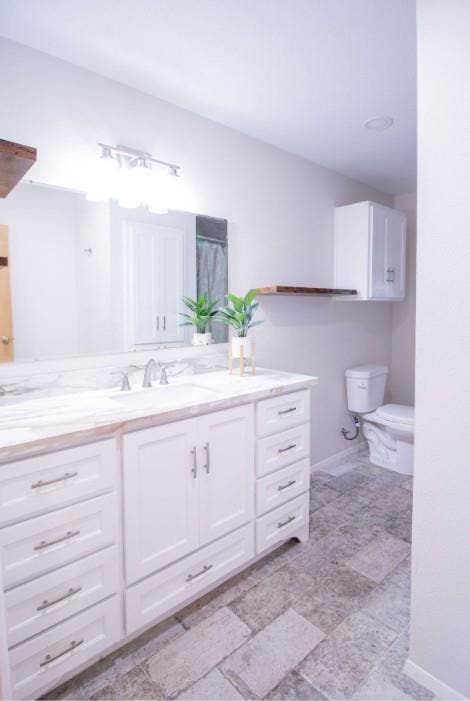 Bathroom featuring a white double-door vanity with stone-finish countertop, undermount sink, multi-drawer storage, and natural stone-finish floor tiling, complemented by white wall-mounted cabinetry and a floating wood-finish shelf