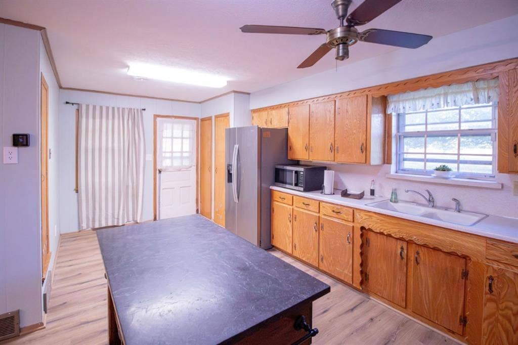 Kitchen featuring wood cabinetry, integrated central island with dark surface, stainless steel refrigerator, white dual-basin sink, and wood-finish flooring