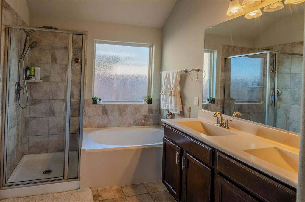 Bathroom featuring a dual-sink vanity with dark-finish cabinetry, a separate soaking tub, a tiled walk-in shower with glass enclosure, and a full-width vanity mirror