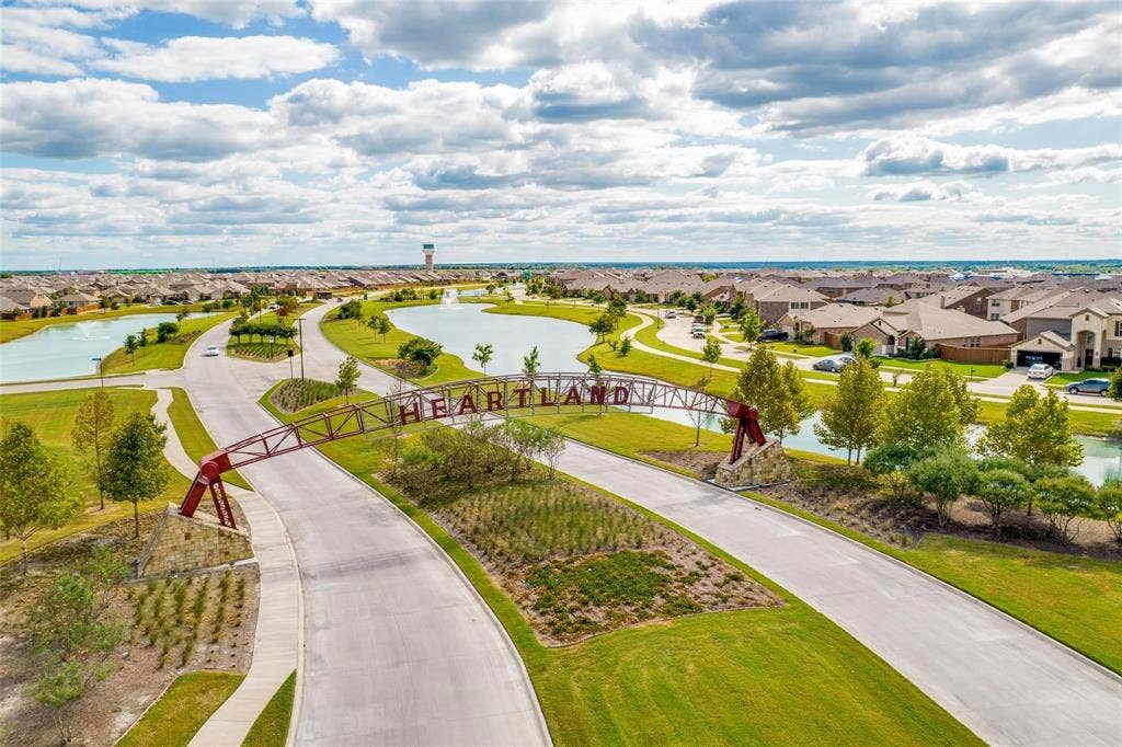 Community entrance featuring a metal arch structure, stone-based supports, ornamental ponds, manicured landscaping, and concrete walkways