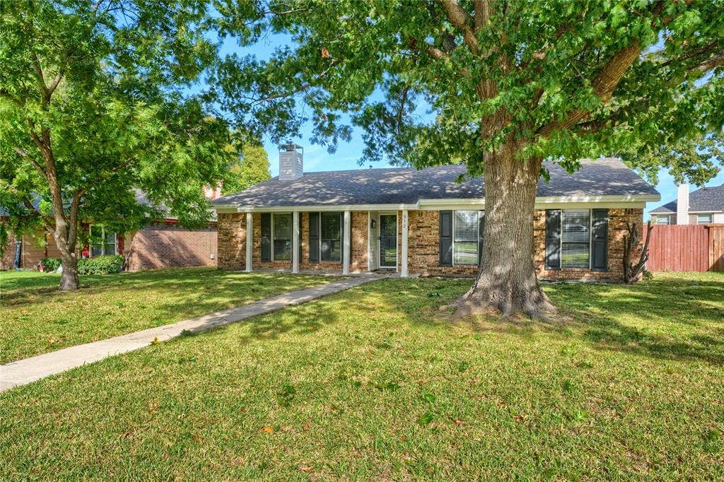 Single-story brick exterior featuring a covered front porch with white columns, shutter-framed windows, and mature trees
