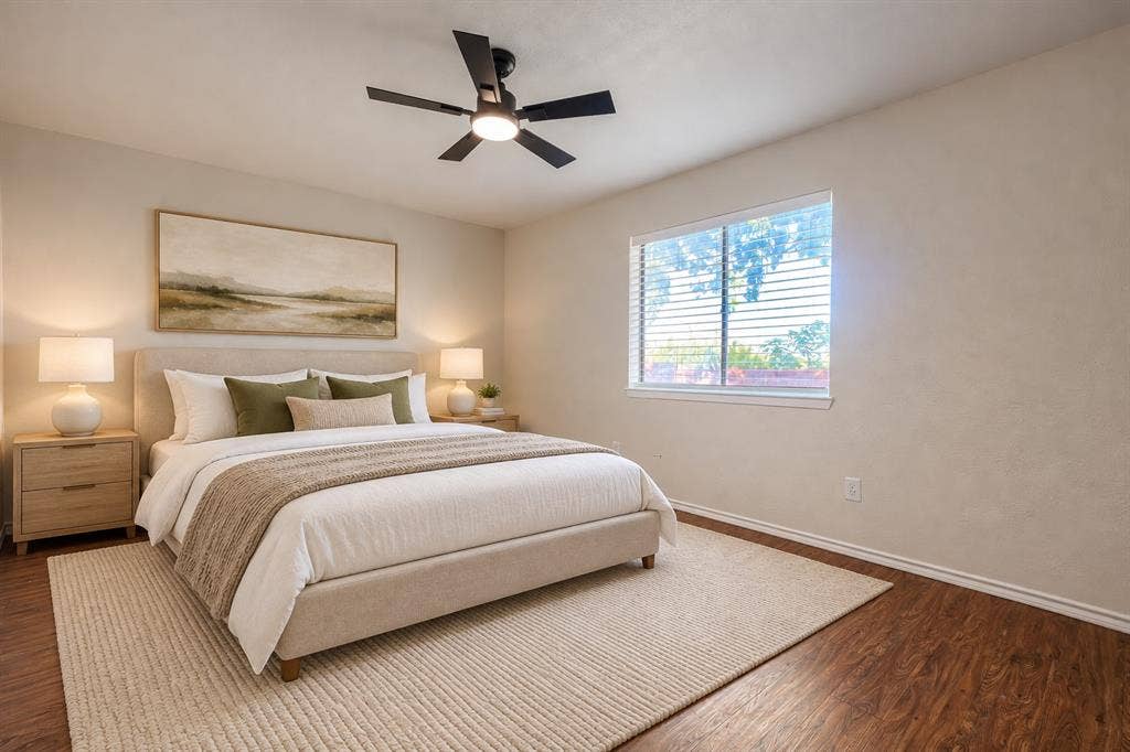 Wood-finish flooring, neutral wall tones, large window with horizontal blinds, and a central ceiling fan with integrated lighting
