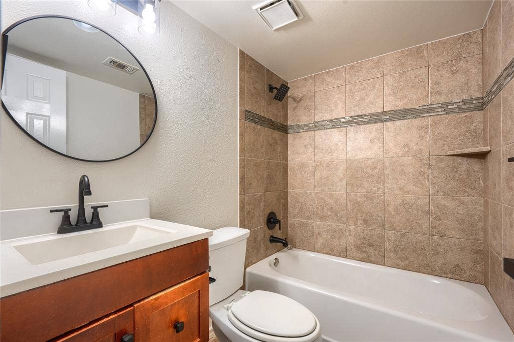Bathroom featuring a wood-finish vanity with a rectangular sink, matte black hardware, a circular mirror, and a tub-shower combination with decorative tile inlay