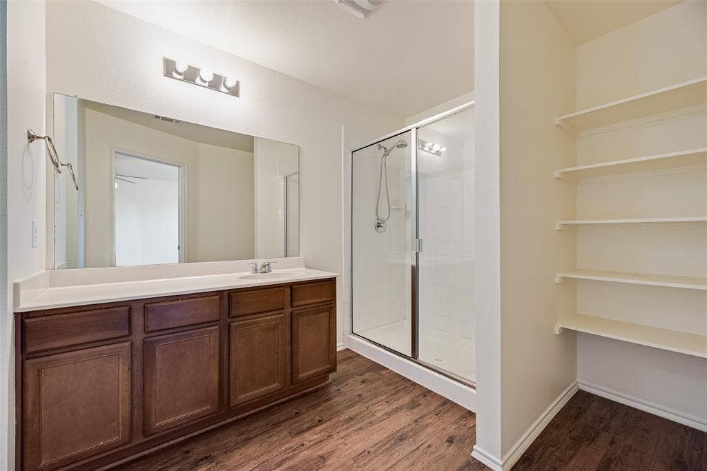 Bathroom featuring a wood-finish vanity with a solid-surface countertop, full-width mirror, walk-in glass-enclosed shower, and built-in shelving