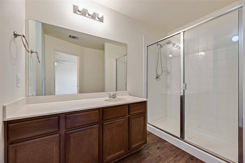 Wood-finish cabinetry with a white solid-surface countertop, full-width vanity mirror, polished chrome lighting fixture, and a glass-enclosed shower with tile surround