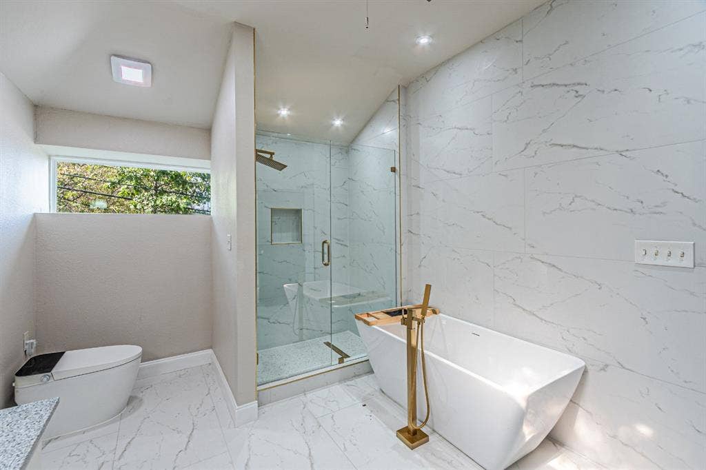 Freestanding soaking tub with a floor-mounted brass faucet, walk-in glass-enclosed shower, built-in shower niche, and large-format tile flooring and wall surfacing