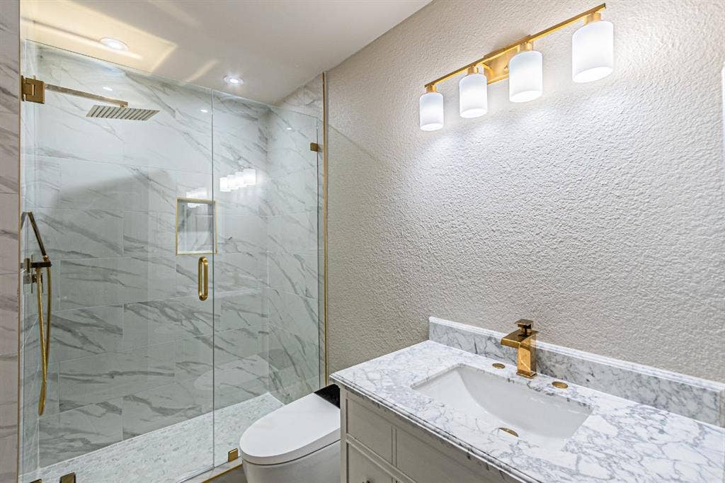 Walk-in shower with floor-to-ceiling large-format tile surround, rainfall showerhead, glass enclosure, gold-tone fixtures, and stone-finish vanity countertop