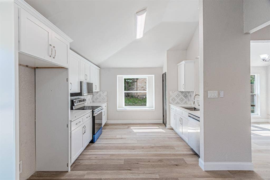 Kitchen featuring white shaker-style cabinetry, wood-finish flooring, patterned tile backsplash, vaulted ceiling, and built-in stainless steel appliances