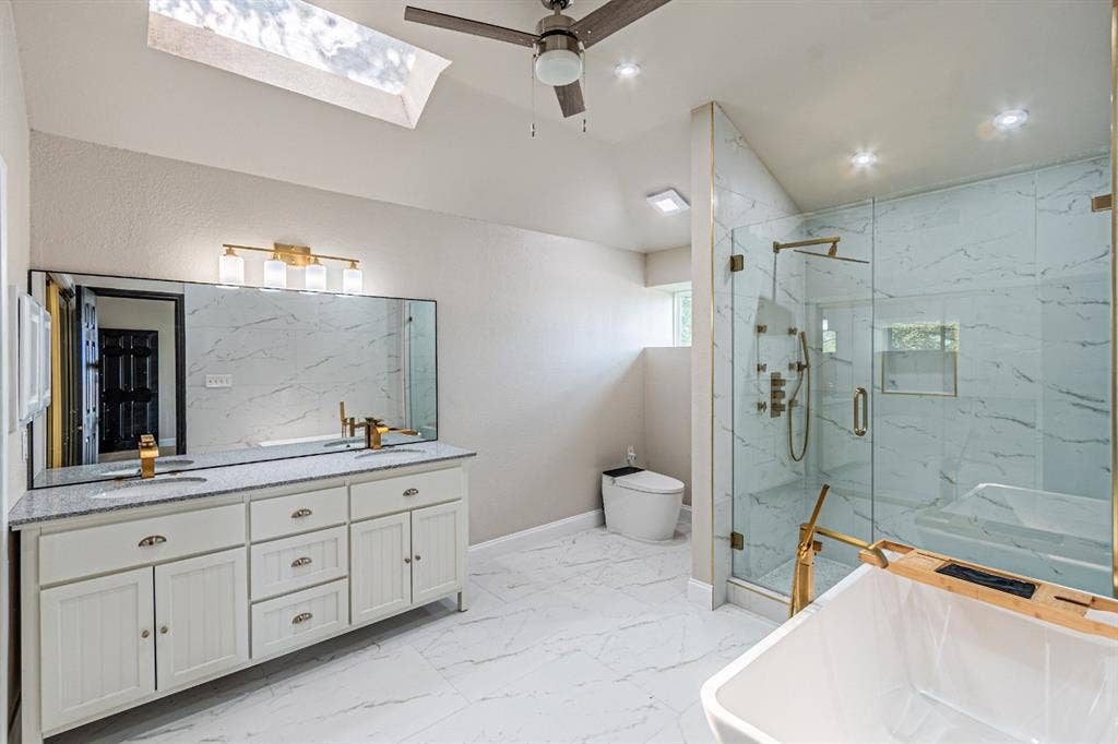 Bathroom featuring a stone-topped double vanity, glass-enclosed walk-in shower with dual shower heads, freestanding tub, skylight, and large-format tile flooring