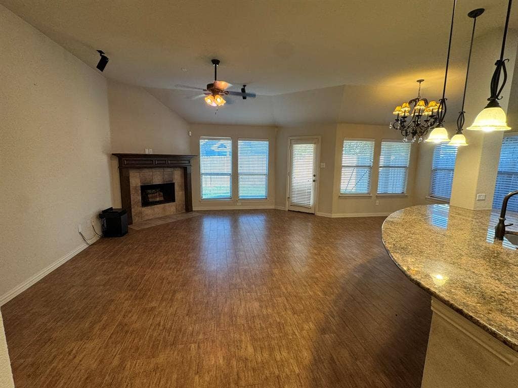 3953 Hollow Lake Road, Fort Worth, TX 76262 Open-concept space featuring wood-finish flooring, vaulted ceilings, a fireplace with stone-surround and timber mantle, stone-surface breakfast bar, and multiple pendant light fixtures